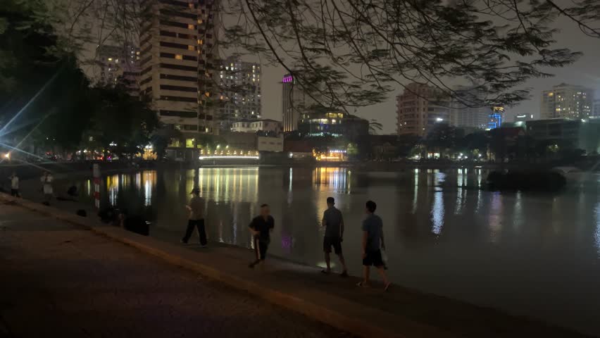 Night view across a city lake in Hanoi, Vietnam: dark water with reflected lights, park foliage on the left and apartment blocks on the far bank.