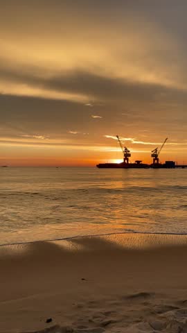Golden sunset over a calm sandy beach with harbor cranes silhouetted against a colorful sky and gentle ocean waves.