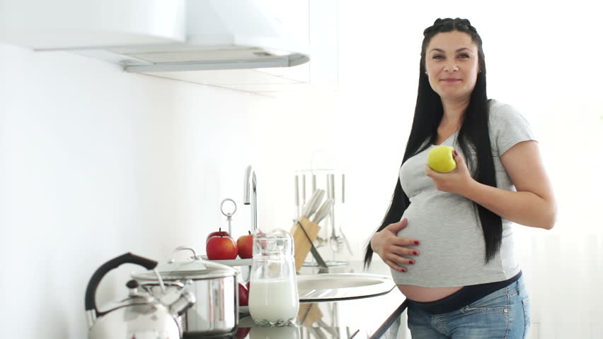 Pregnant woman eating an apple in the kitchen and stroking tummy
