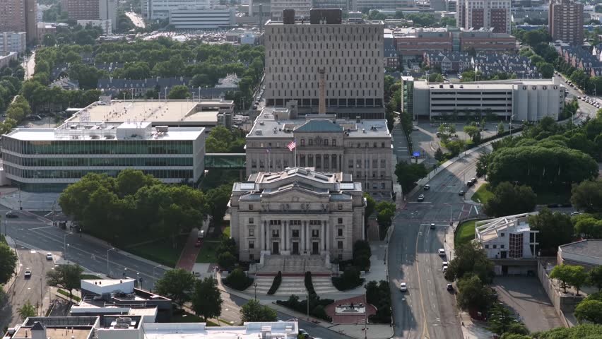 Domed courthouse with classical columns anchors downtown Newark plaza, glass office building and parking garage framing wide tree-lined boulevard with passing traffic. Aerial drone approaching shot