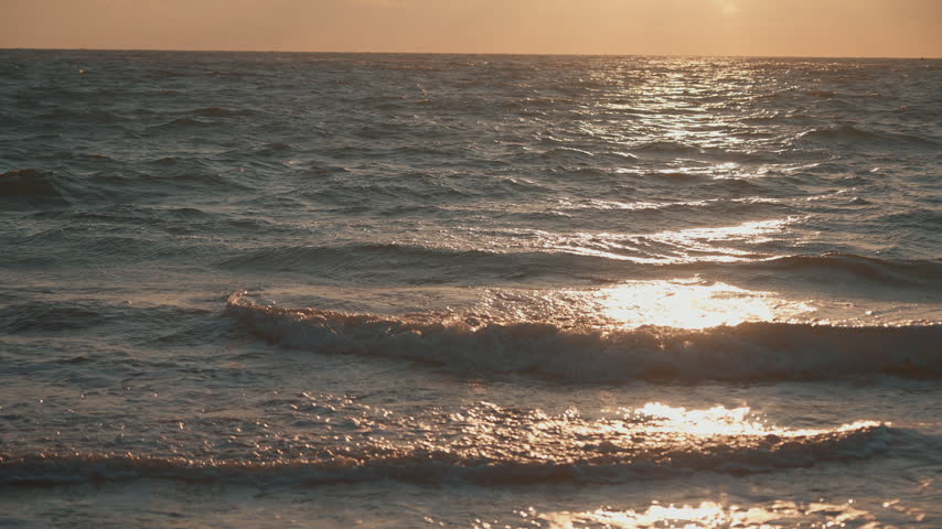 Man walking along the beach by the sea at sunset, approaching the camera and creating a natural foreground transition.