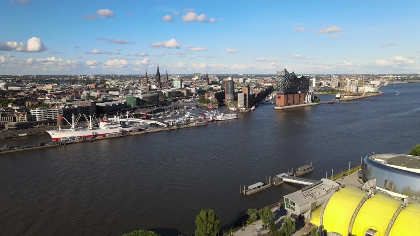 Aerial drone shot overlooking the Elbphilharmonie beside the Elbe River, with ships, docks, waterfront buildings and Hamburg city skyline under a blue sky in Germany
