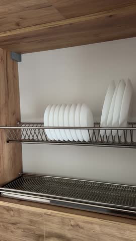 Open kitchen cabinet with stainless steel dish drying rack and neatly arranged white ceramic plates. Modern storage solution for organized kitchen interior.