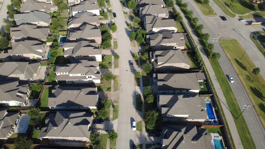 Straight residential avenue extends beside multi lane highway in Dallas Fort Worth metroplex. Aerial shot two story brick homes, dark shingle roofs, backyard fencing, manicured lawns, Frisco, TX. USA