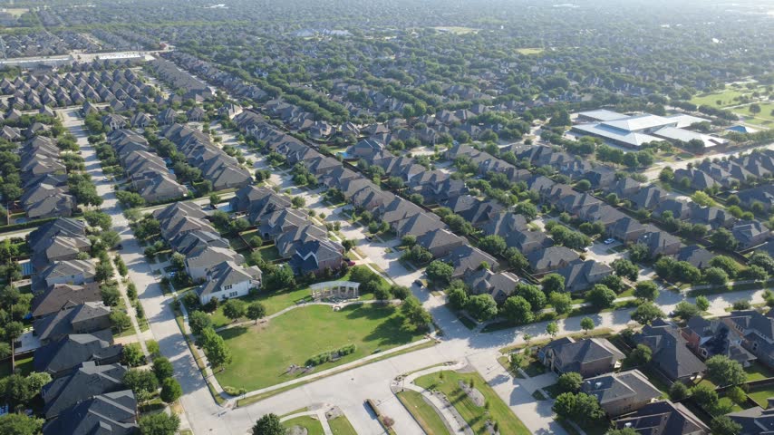 Sprawling public school building borders dense residential blocks in Denton County. High drone angle captures two story brick houses, dark shingle roofs, backyard pools, park lawns, Frisco, TX. USA