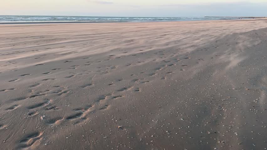 A fine sandy beach where footprints leading to the shore are still visible, with a horizontal sandstorm blowing under a serene sky. The coastline stretches as far as the eye can see, creating a peaceful coastal atmosphere and highlighting the natural patterns etched by footsteps and the wind.