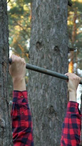 Young man in a plaid shirt grimacing with effort while doing pull ups on a horizontal bar attached to trees in a forest, engaging in an intense outdoor workout to build muscle