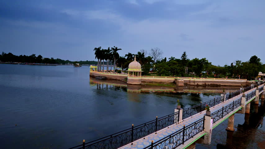 Stone paths lead across peaceful waters toward island palm trees. Heavy monsoon clouds cast dramatic reflections. Picturesque scenery stretches through Udaipur, Rajasthan.