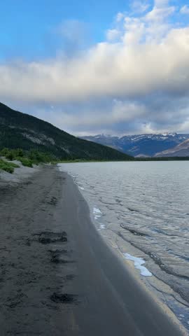 Vertical landscape featuring a wide sandy beach shoreline along the rippled water of Talbot Lake with the sunlit peaks of the Colin Range under a blue sky with clouds in Jasper National Park, Alberta, Canada.