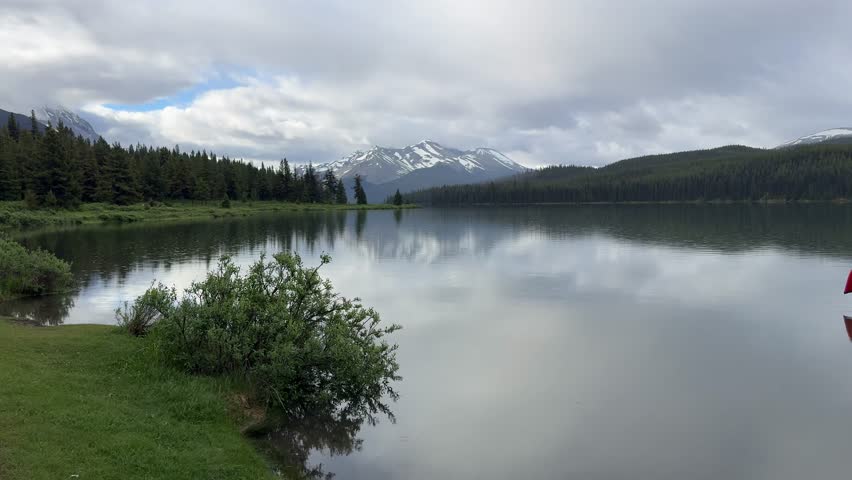 Landscape featuring stacked red canoes on a wooden dock with calm water reflections of mountains and forest under a cloudy sky at Maligne Lake in Jasper National Park, Alberta, Canada.