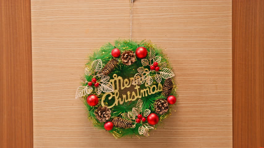 A man reaches out and adjusts a green Christmas wreath decorated with red baubles, pinecones and gold lettering hanging on a wooden door.