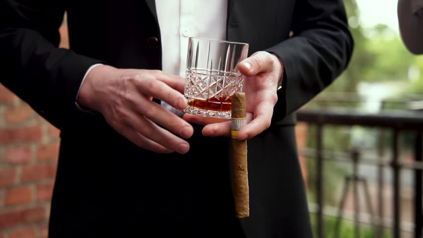 Businessman in a luxury suit holding a crystal glass of whiskey and a premium cuban cigar close-up.