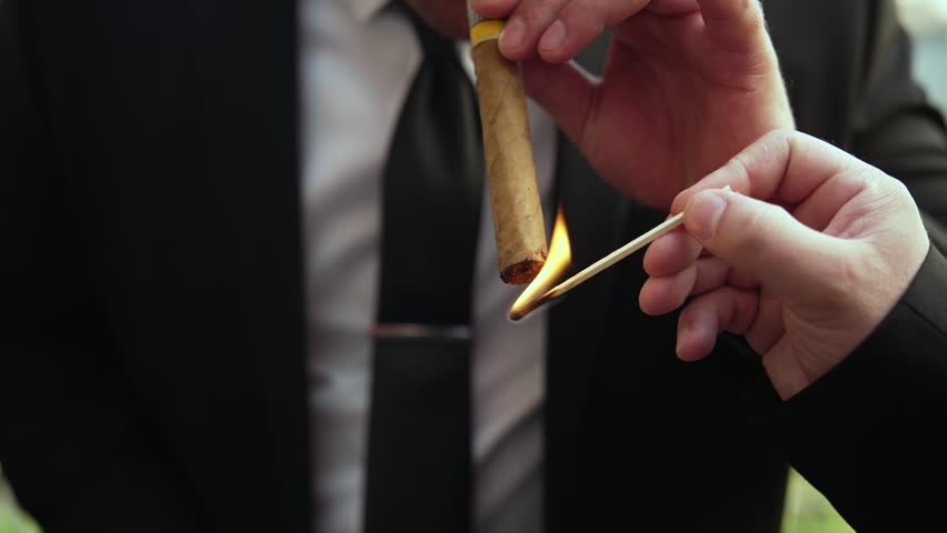 Man in a business suit holding a premium cuban cigar and lighting it with a wooden match close-up.