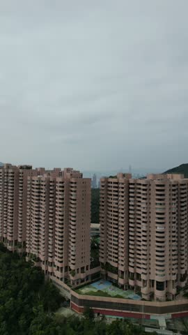 Aerial view moving away from residential buildings in Hong Kong, revealing the vast city skyline with its modern skyscrapers and green mountainsides under a cloudy and overcast sky.