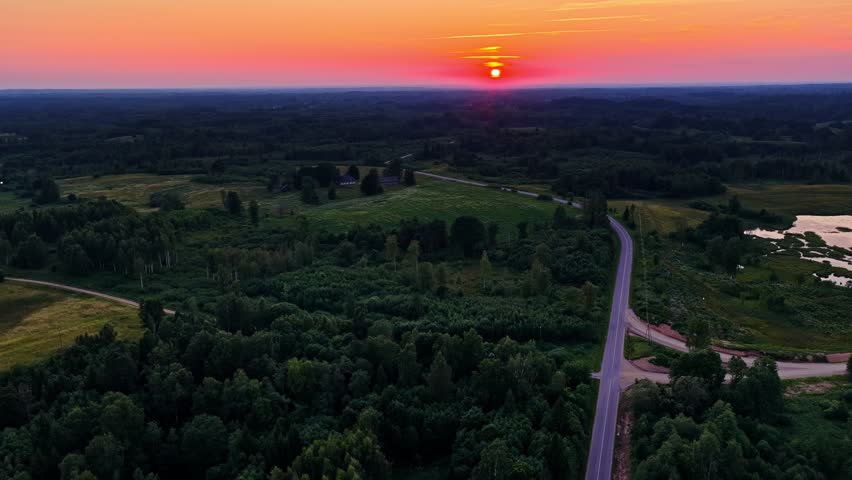 Bright sunset descending over a lush green forest, meadows, and a winding rural road at dusk