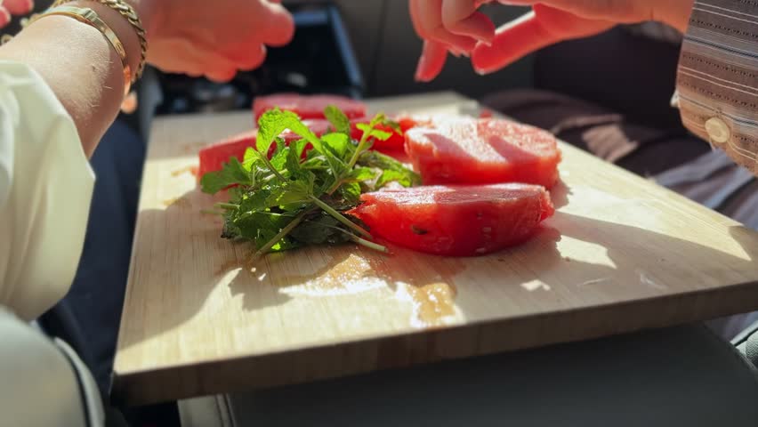 Close up shot of hands taking fresh sliced watermelon and green mint leaves from a wooden cutting board. The healthy summer snack is shared inside a sunny car interior during a road trip.