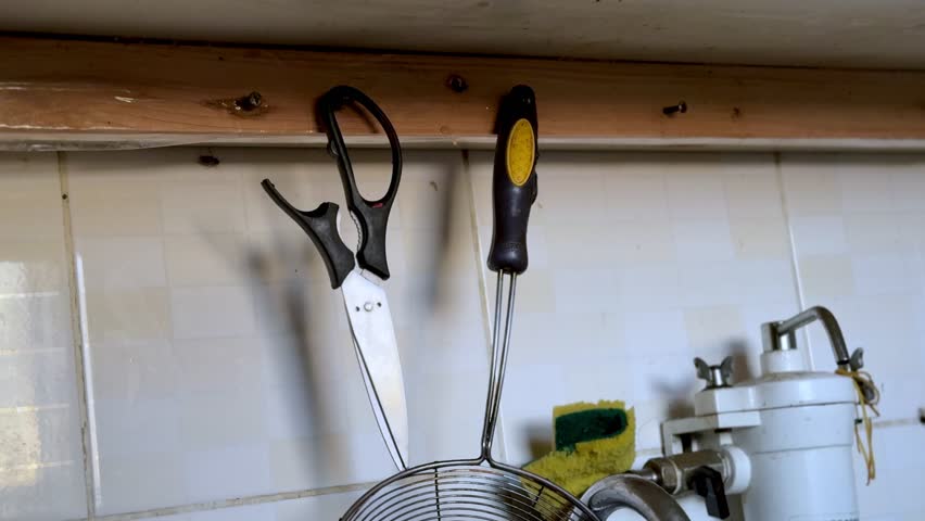 A close-up static shot of kitchen scissors and a metal strainer hanging on a wooden beam. The cooking tools rest against a white tiled wall near a sink and sponge in a domestic indoor kitchen.