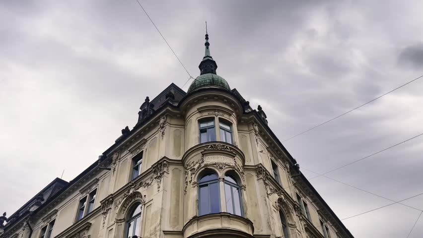 Low-angle view of an ornate historic corner building in Ljubljana, featuring a rounded bay, arched windows, sculpted facade and copper-domed turret beneath dramatic clouds.