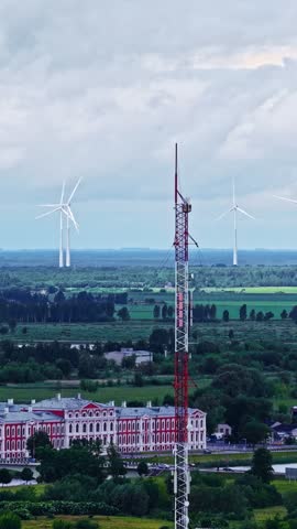 Tall red and white communication tower stands above a rural landscape with spinning wind turbines on a cloudy day.