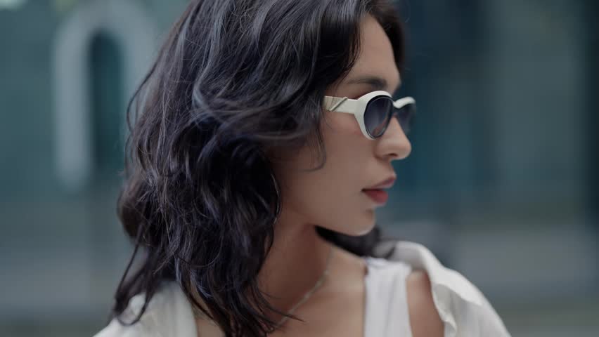 Confident beautiful woman with dark wavy hair wearing trendy sunglasses turning her head and looking at the camera in slow motion. Portrait of a serious attractive girl posing outdoors