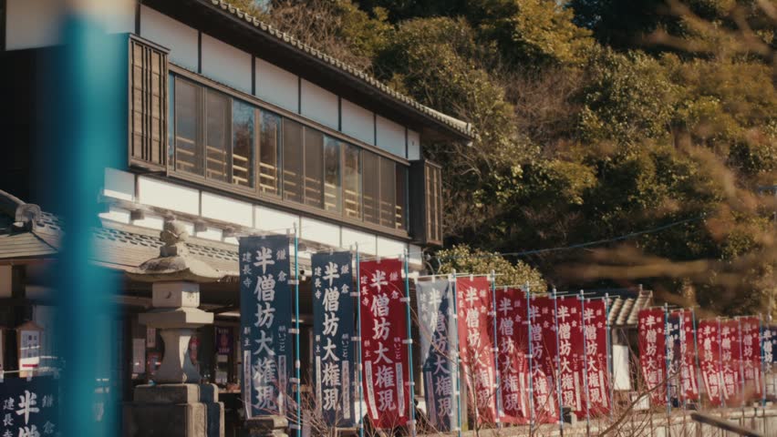 The traditional wooden shrine building of Hansobo, located atop the Kencho-ji temple complex in Kamakura, framed by rows of ceremonial banners and wooded hillside, with warm autumn light on the trees.