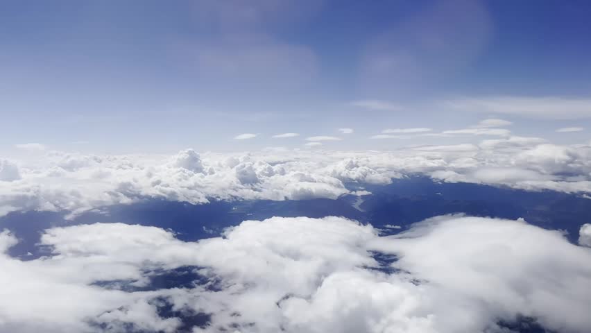 Airplane window view above cloud layers and distant alpine mountains, with blue sky, white clouds and hazy high altitude travel scenery.