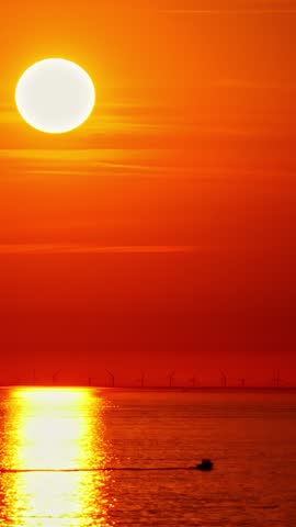 Huge orange sun sets over calm sea as a distant wind farm and a small speedboat silhouette pass through the glowing reflection.