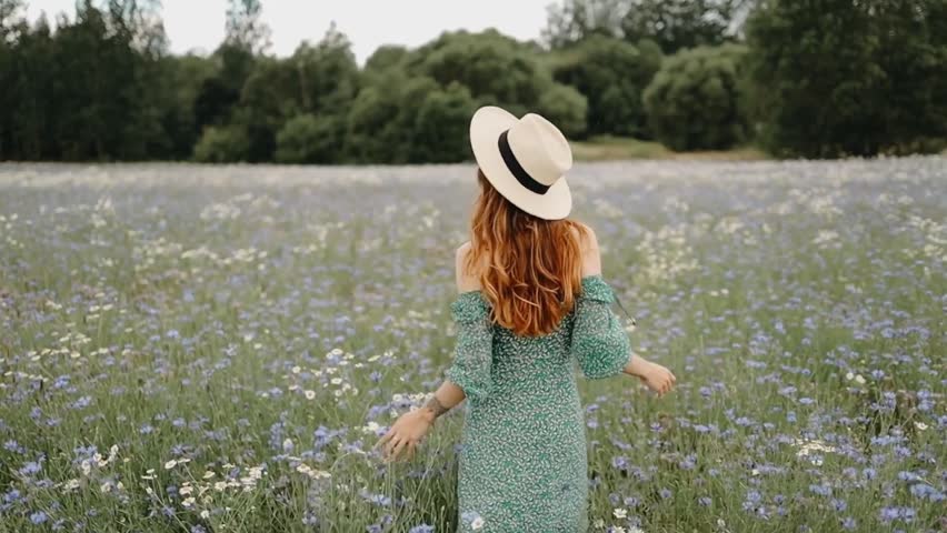 Rear view of a young woman with long wavy auburn hair wearing a cream straw fedora hat with black band and a green floral off-shoulder dress, walking through a lush meadow filled with blue cornflowers and white wildflowers. Tall green trees form a natural backdrop under a soft overcast sky, creating a dreamy, romantic countryside atmosphere. Perfect for lifestyle, travel, summer, and nature-themed content.