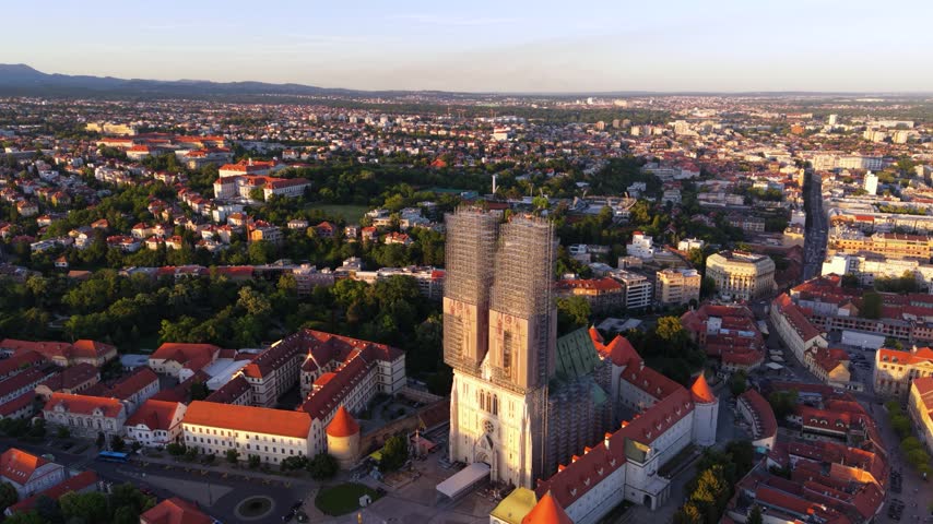 Aerial view of Zagreb Cathedral undergoing restoration with extensive scaffolding around its twin towers, surrounded by historic red-roofed buildings and the Zagreb cityscape in warm golden-hour light.