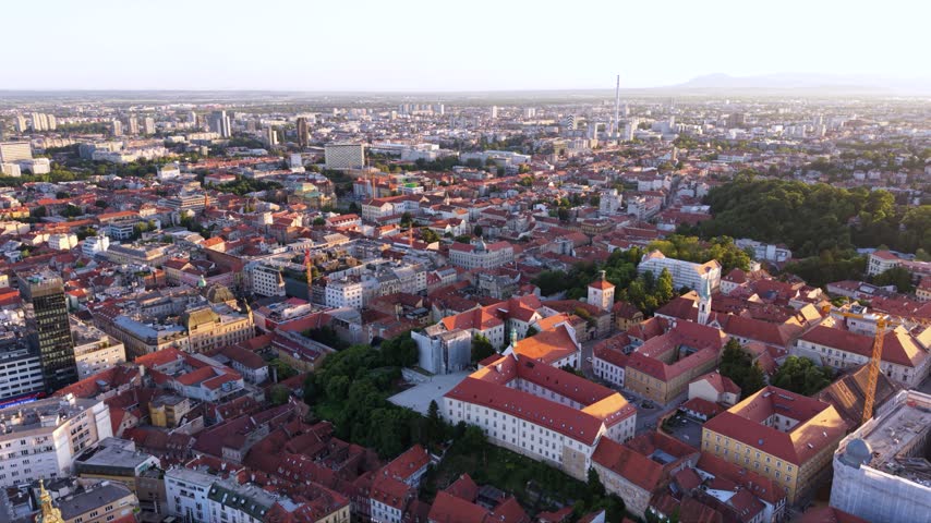 Aerial view of Zagreb Cathedral undergoing restoration with extensive scaffolding around its twin towers, surrounded by historic red-roofed buildings and the Zagreb cityscape in warm golden-hour light.