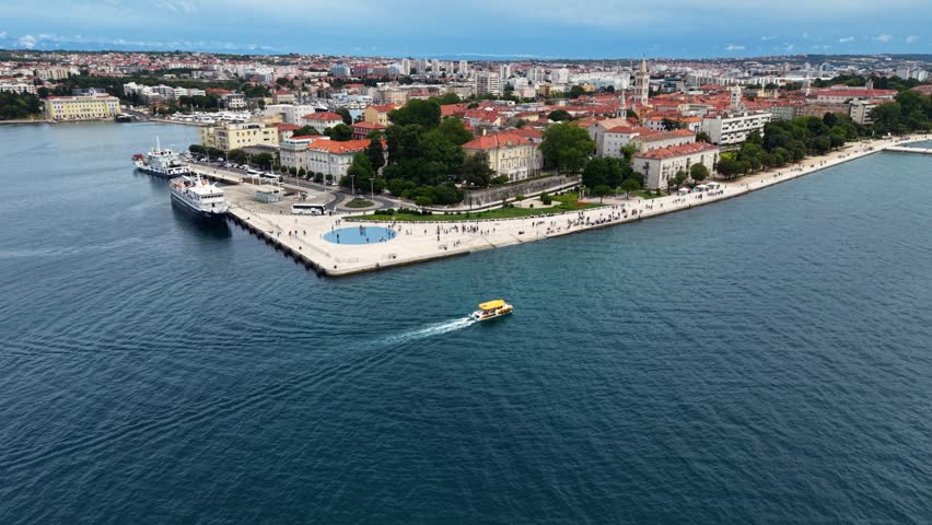 Wide aerial view of Zadar, Croatia, showing the historic old town peninsula, marina, waterfront and expansive Adriatic Sea, with a small passenger boat crossing the bay.