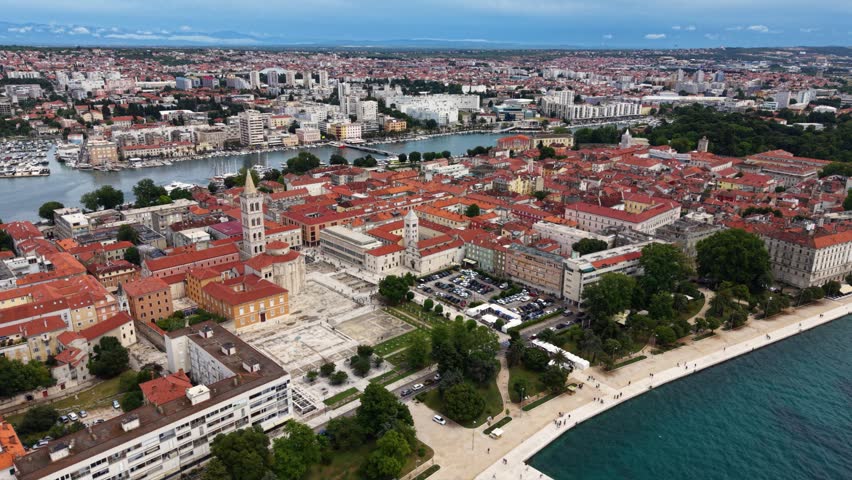 Aerial view of the historic center of Zadar, Croatia, showing red tiled rooftops, church towers, Roman Forum ruins, waterfront promenade, marina and the surrounding modern city beside the Adriatic Sea.