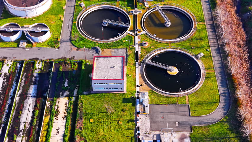 Drone view of circular wastewater clarifier. Rotating skimmer arm moves across dark water tank beside a red roofed control building and green lawn.