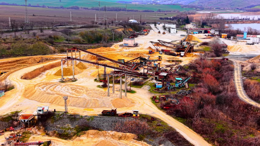 Overcast view of quarry complex and surrounding fields. Aerial perspective shows conveyors, stockpiles, power lines, and distant mountains.