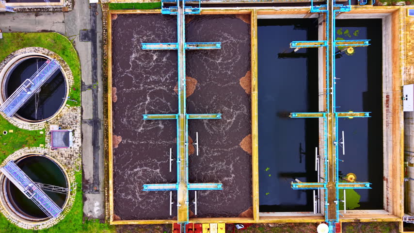 Overhead view of aeration basins at wastewater plant. Bird eye angle showing frothy brown water beside clear settling tank with blue walkway bridges.