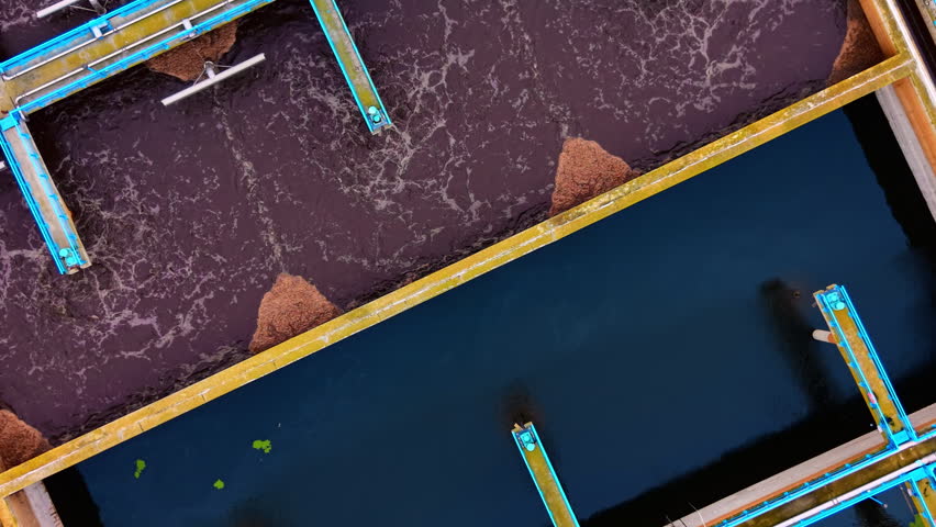 Aerial close up of frothy wastewater treatment pool. Overhead detail of foamy brown aeration basin beside dark clarifier tank with metal walkway.