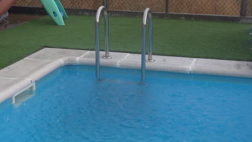 Raindrops falling into a swimming pool on a stormy day. The downpour creates ripples and splashes on the surface of the blue water