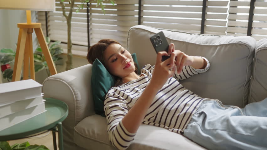 Overhead view of relaxed young Asian woman lying on sofa smiling using smartphone, cozy home living room with plants and side table, top-down casual lifestyle scene with copy space