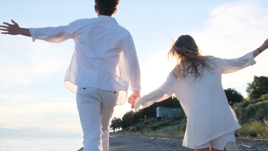 Back view, young couple in white clothes run along the seashore at sunset with arms spread. Romantic date, youthful love, beach romance, happy relationship and joyful moments