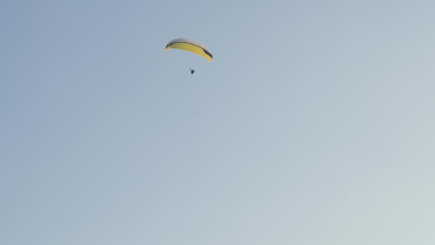 Parachute glides through clear blue sky. One person floats above, suspended in calm flight. Sunlight glows softly behind distant canopy. Ideal for adventure, freedom, or tranquil travel themes