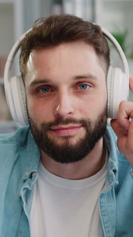 Headshot close up face portrait of Caucasian good-looking man looking and posing at camera while listen to music in white headphones in living room at home, inside