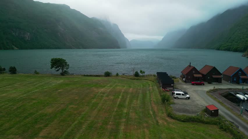 Scenic fjord landscape featuring colorful boathouses and parked boats along the shoreline, with lush mountains and a cloudy sky in the background
