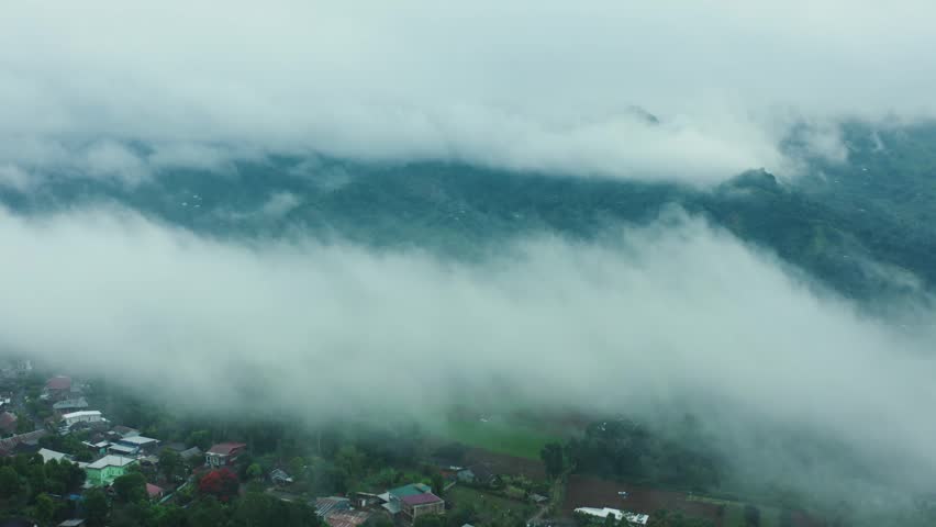 Aerial drone cinematic view of a quiet village partially veiled by thick rolling clouds and heavy morning fog over mountain ranges in 4K resolution.