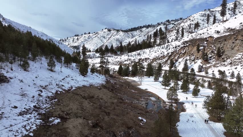 Aerial drone footage captures a scenic winter landscape featuring the Kirkham Hot Springs with visitors walking along a snowy path beside a river and a winding mountain road