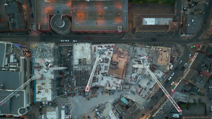 An aerial view at dusk captures a vast city construction site with multiple cranes, a prominent spiral car park, and surrounding urban streets illuminated by evening lights and traffic.
