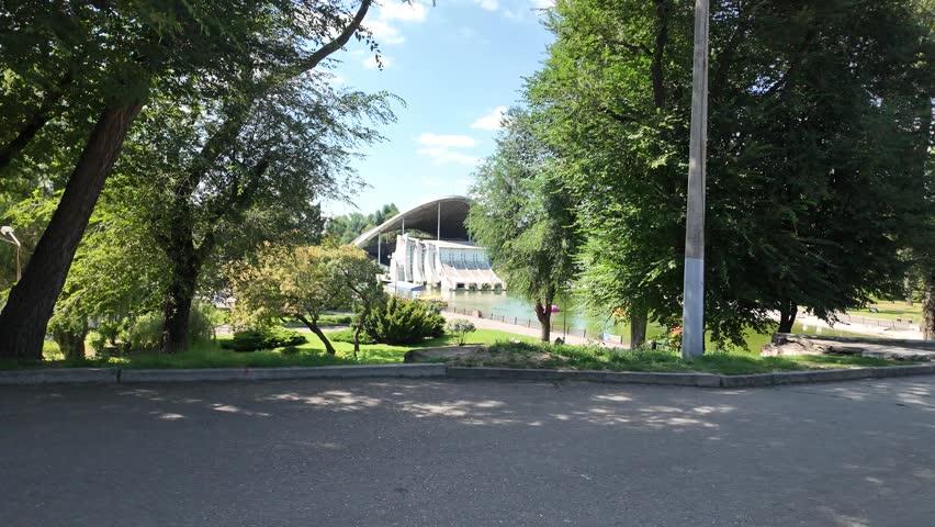 Picturesque city park landscape featuring a pond, pedestrian walkway along the water, green trees, and an architectural amphitheater stage under a clear blue sky.
