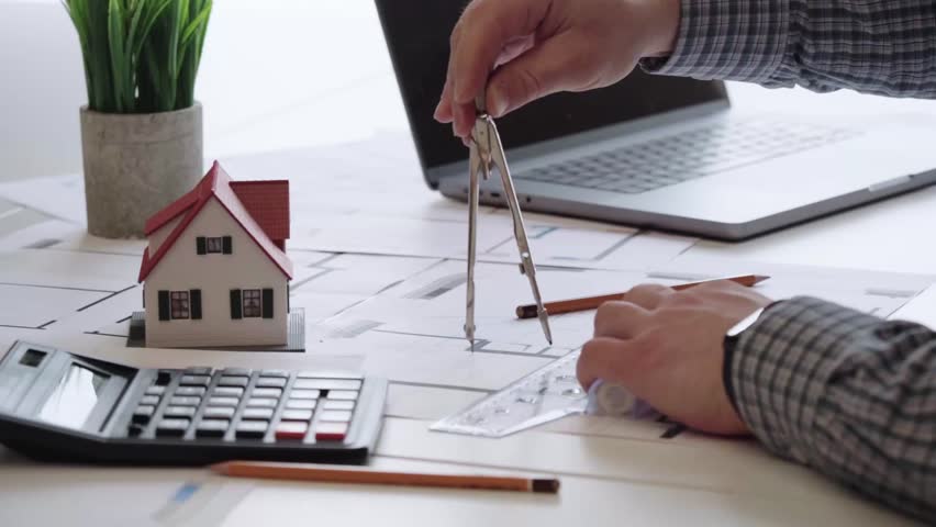 Hand draws plans with compass near miniature home. Calculator and laptop sit beside the workspace. Desk holds blueprint and small house model. Professional mood, focused on design and budgeting