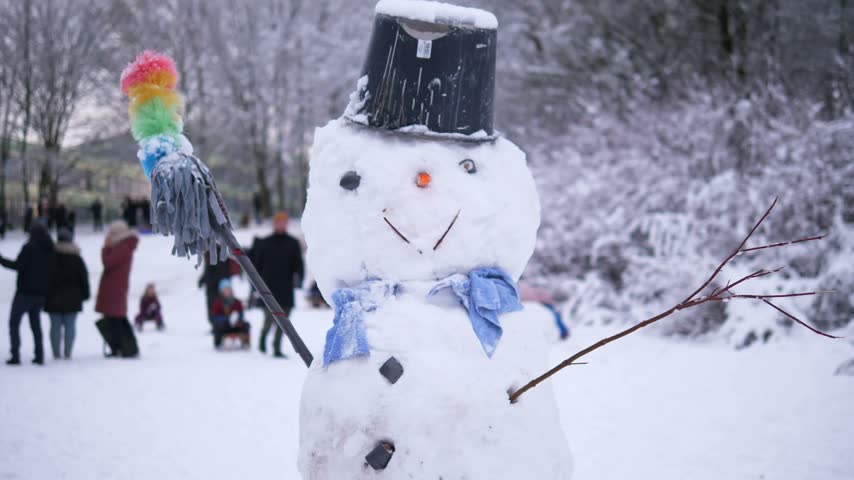 Snowman with a black hat, blue scarf and colorful broom standing in a snowy winter park with people in the background
