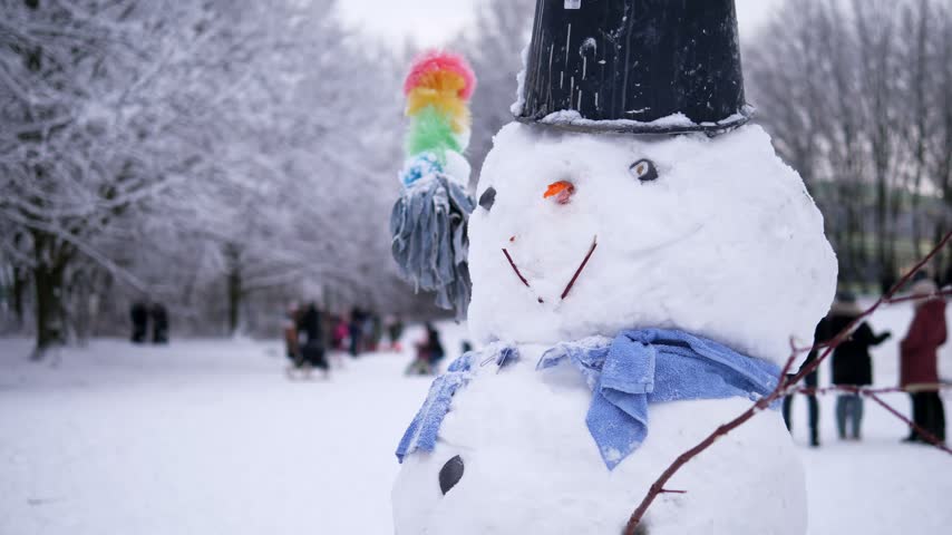Snowman with a black hat, blue scarf and broom standing in a snowy winter park with people in the background. The concept of winter and Christmas