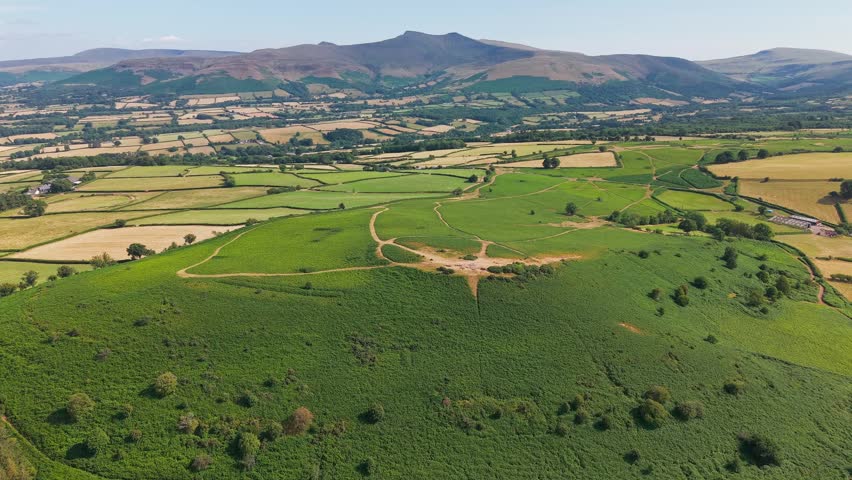Grassy ridge of an ancient hillfort surrounded by dry summer farmland in Wales, clear blue skies and bright sunlight highlighting ancient tracks and field boundaries